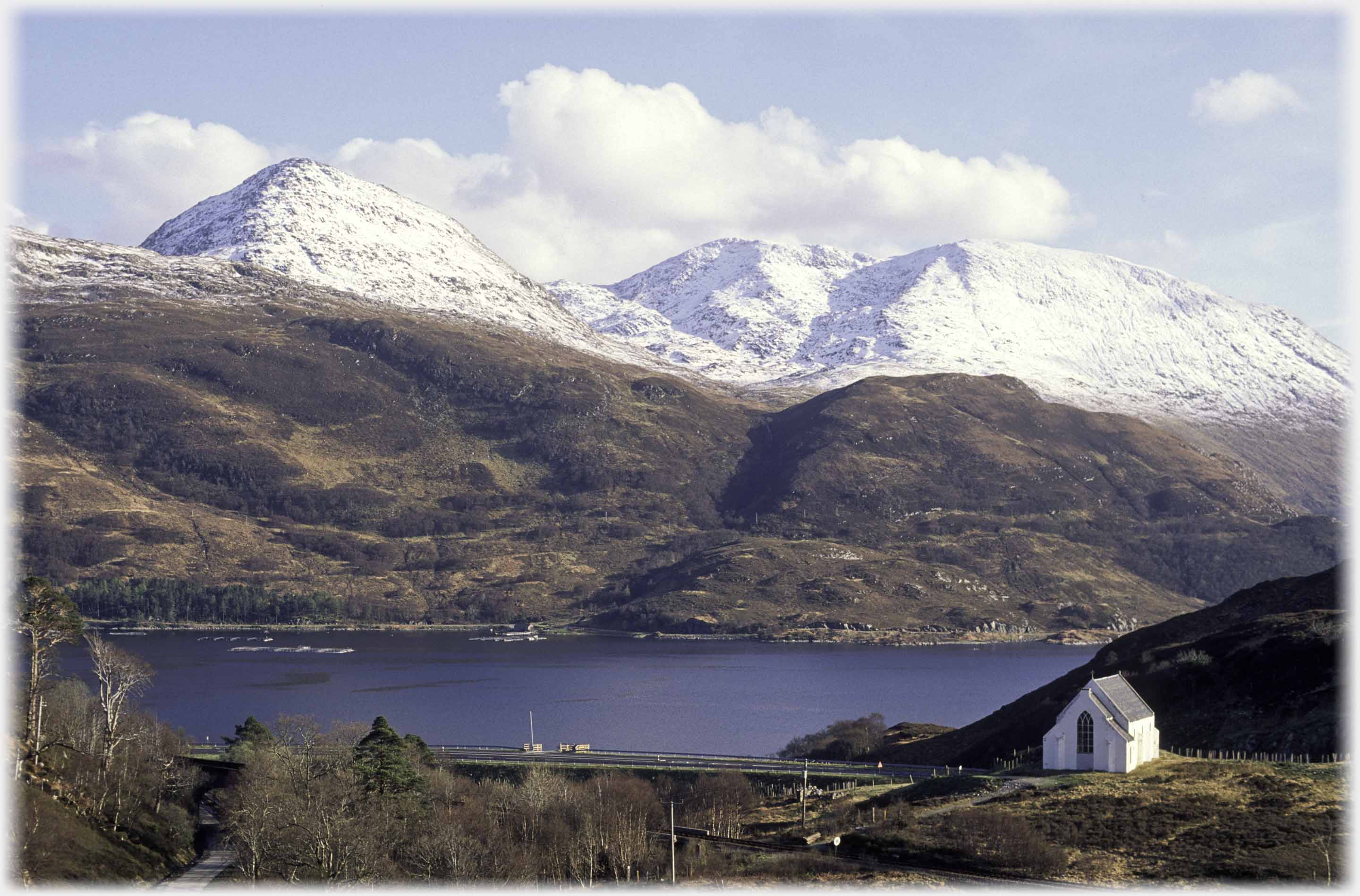 Small white church with water beyond and snow covered hills behind water.