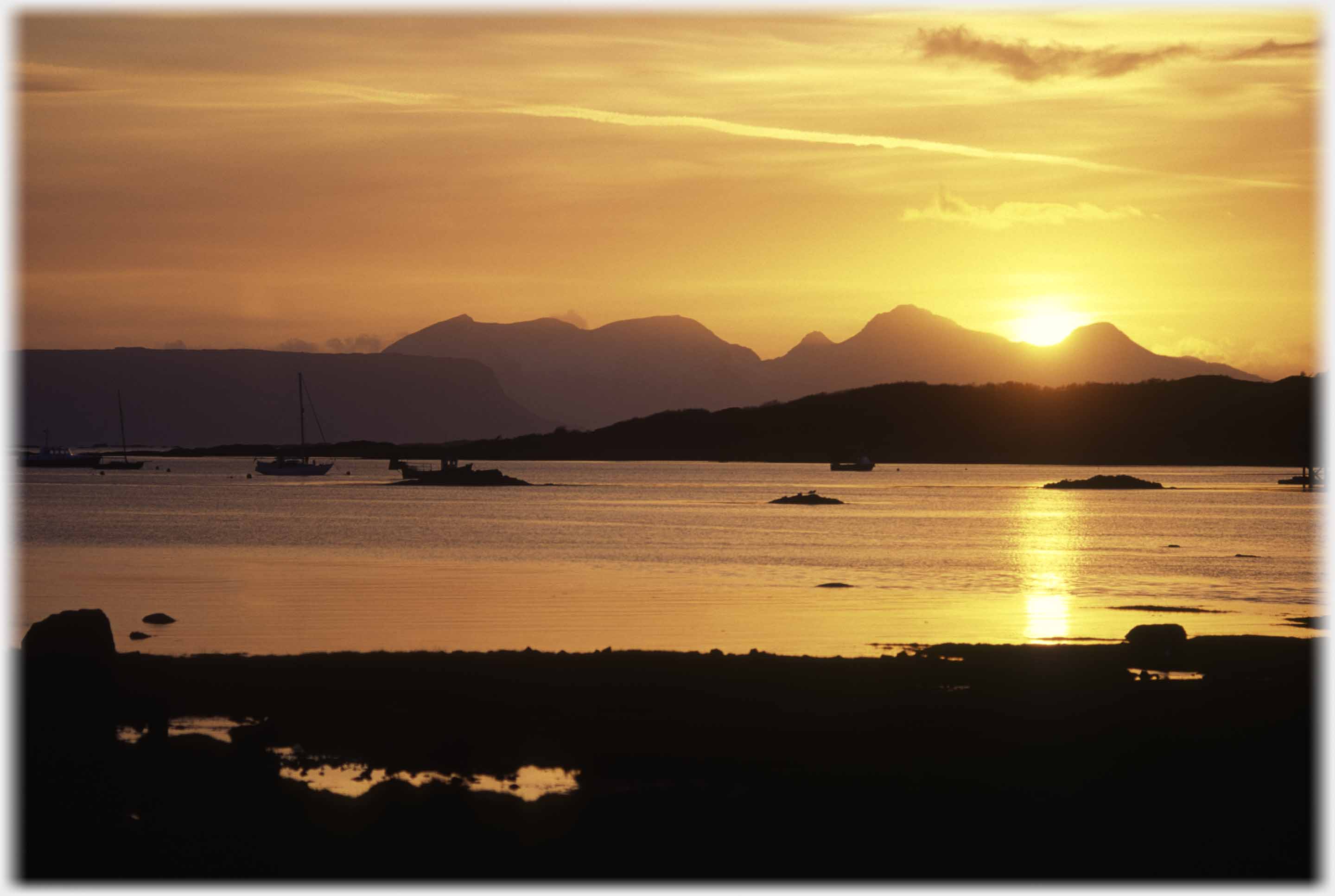 Golden sunset with sun touching mountains and reflected in sea.