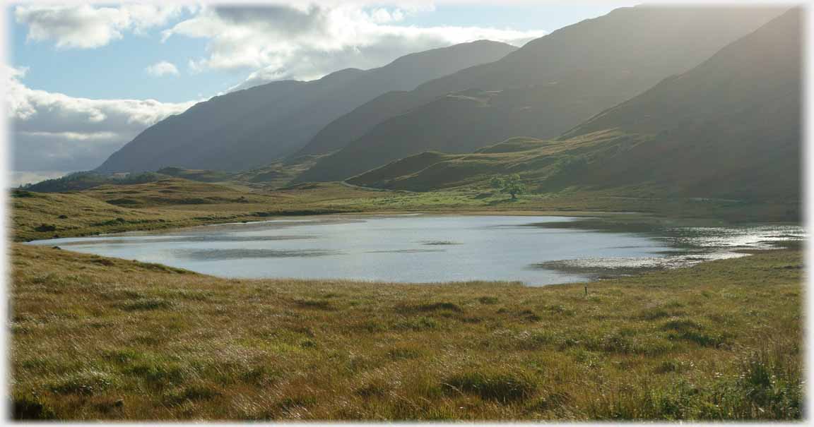 Lochan seen into the sun with hills runniing down to it.