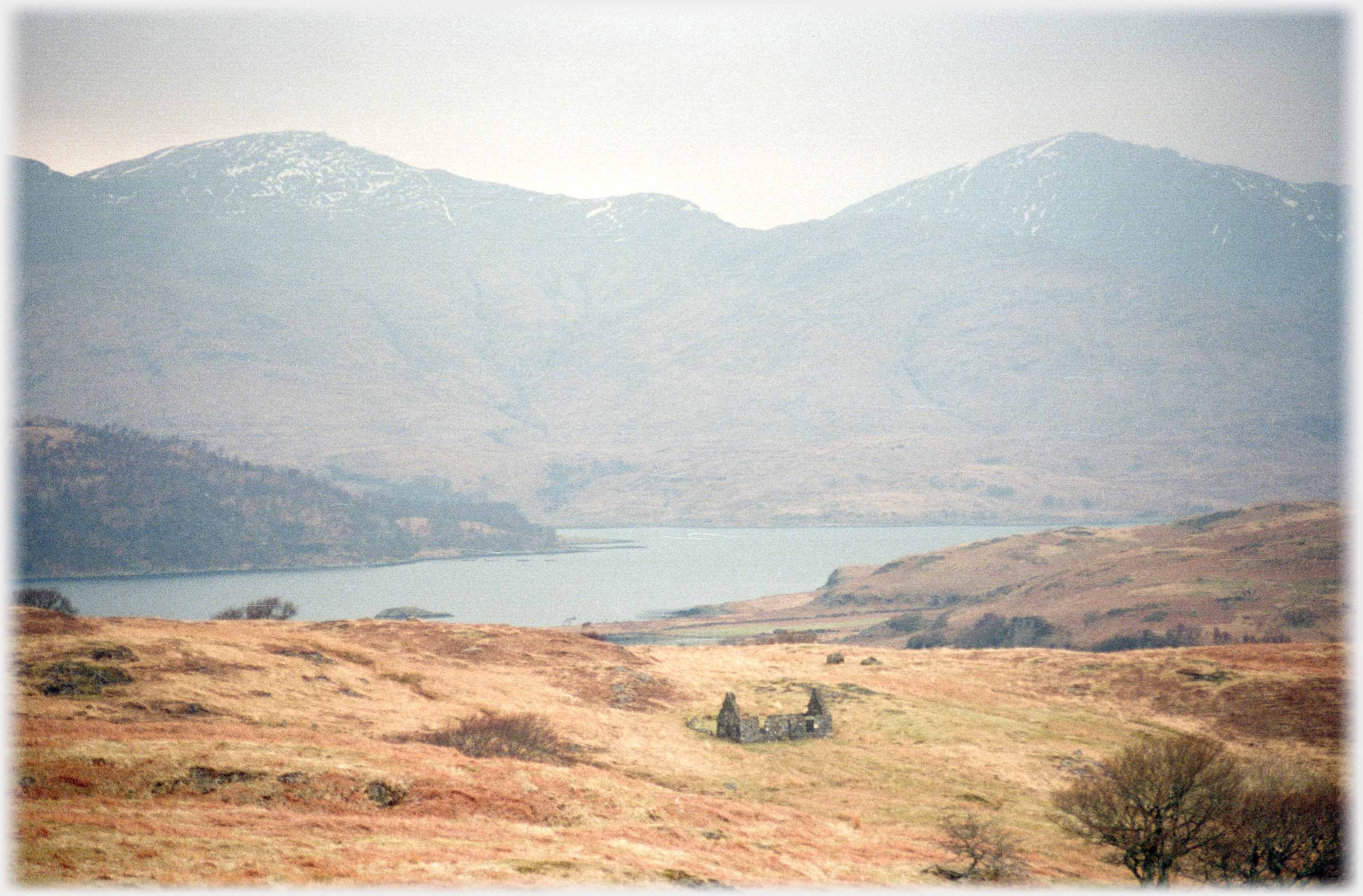 Wide shot of loch with hills beyond and ruined cottage on moorland in foreground.