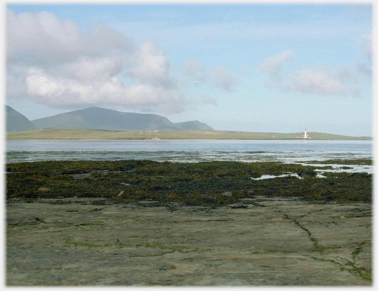 Foreground shore with hills in distance and lighthouse across the water.
