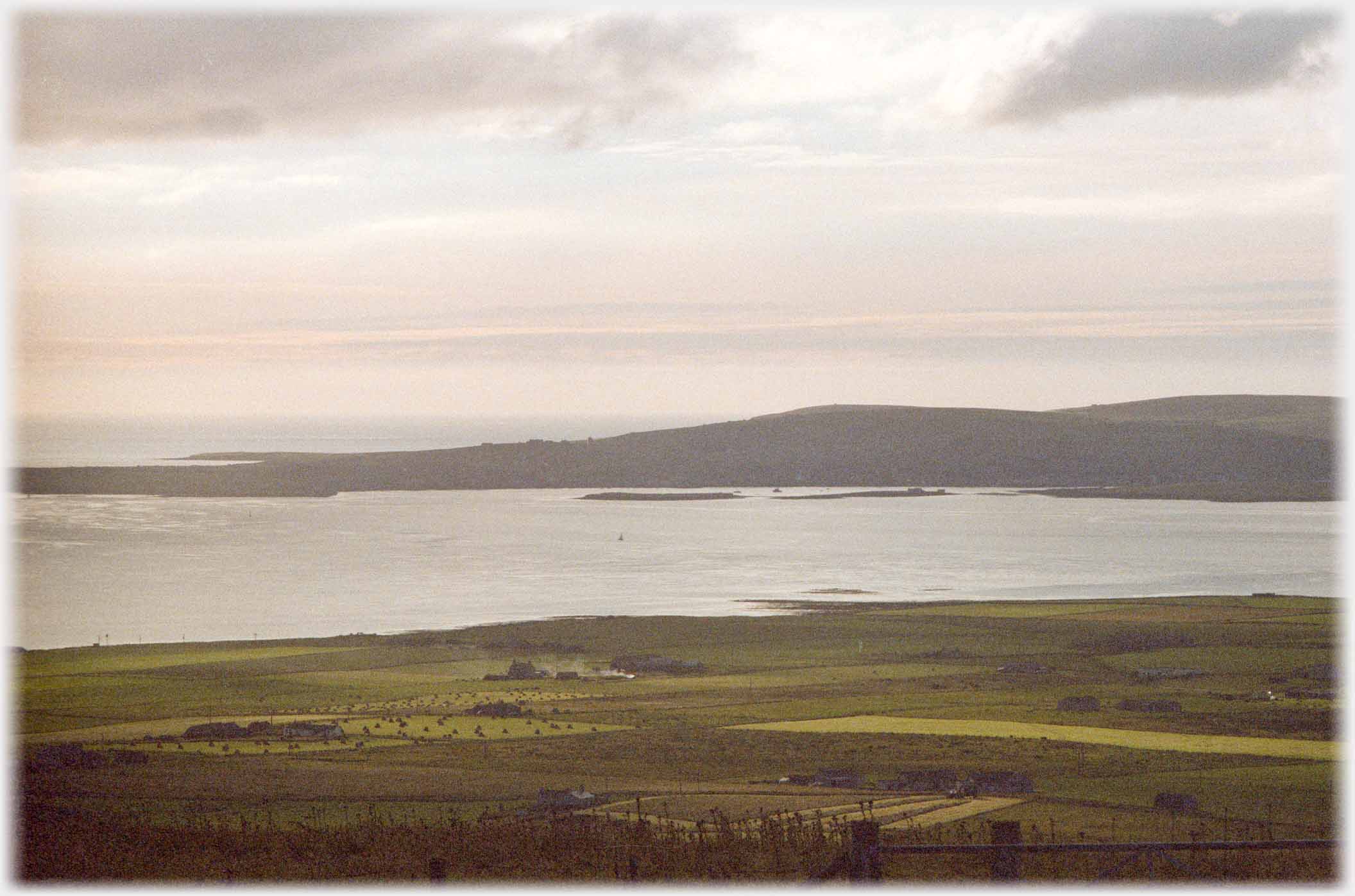 Foreground of fields, with land beyond  water and islets in evening light.