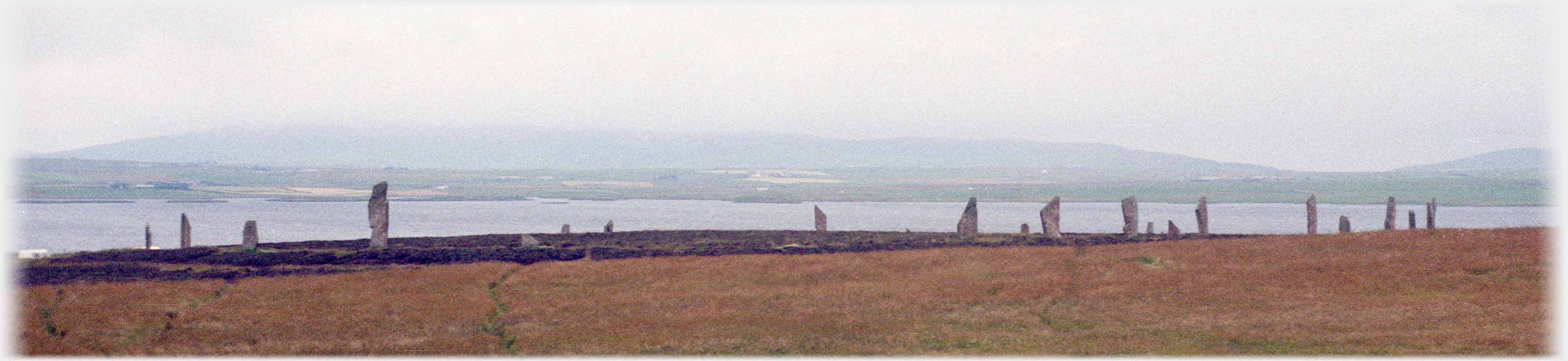 Large stone circle on near horizon, sea and hill beyond.