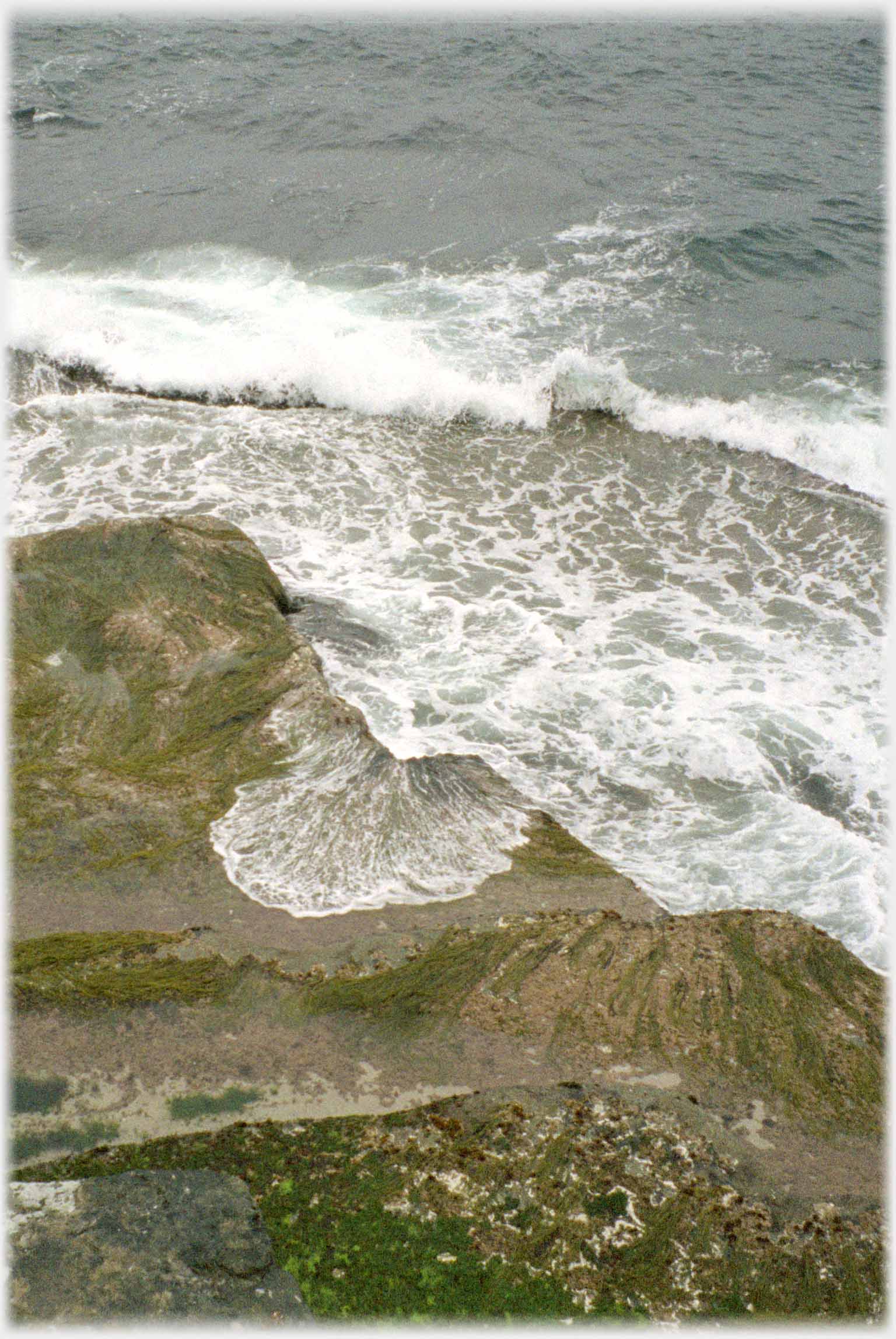 Looking down on breakers forming patterns of foam on the cliff foot rocks.
