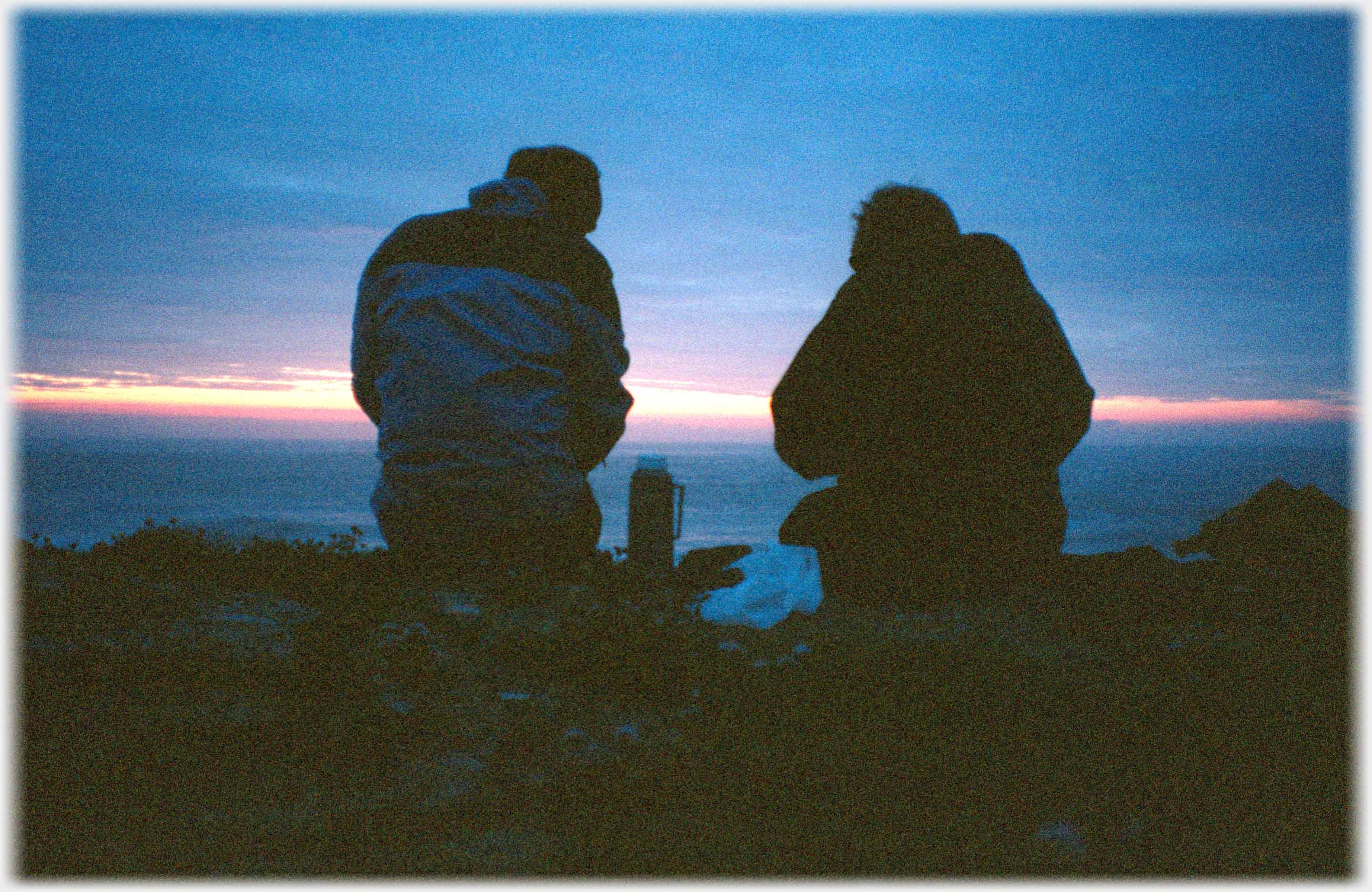 Two silhouettes and flask against last of light over sea.