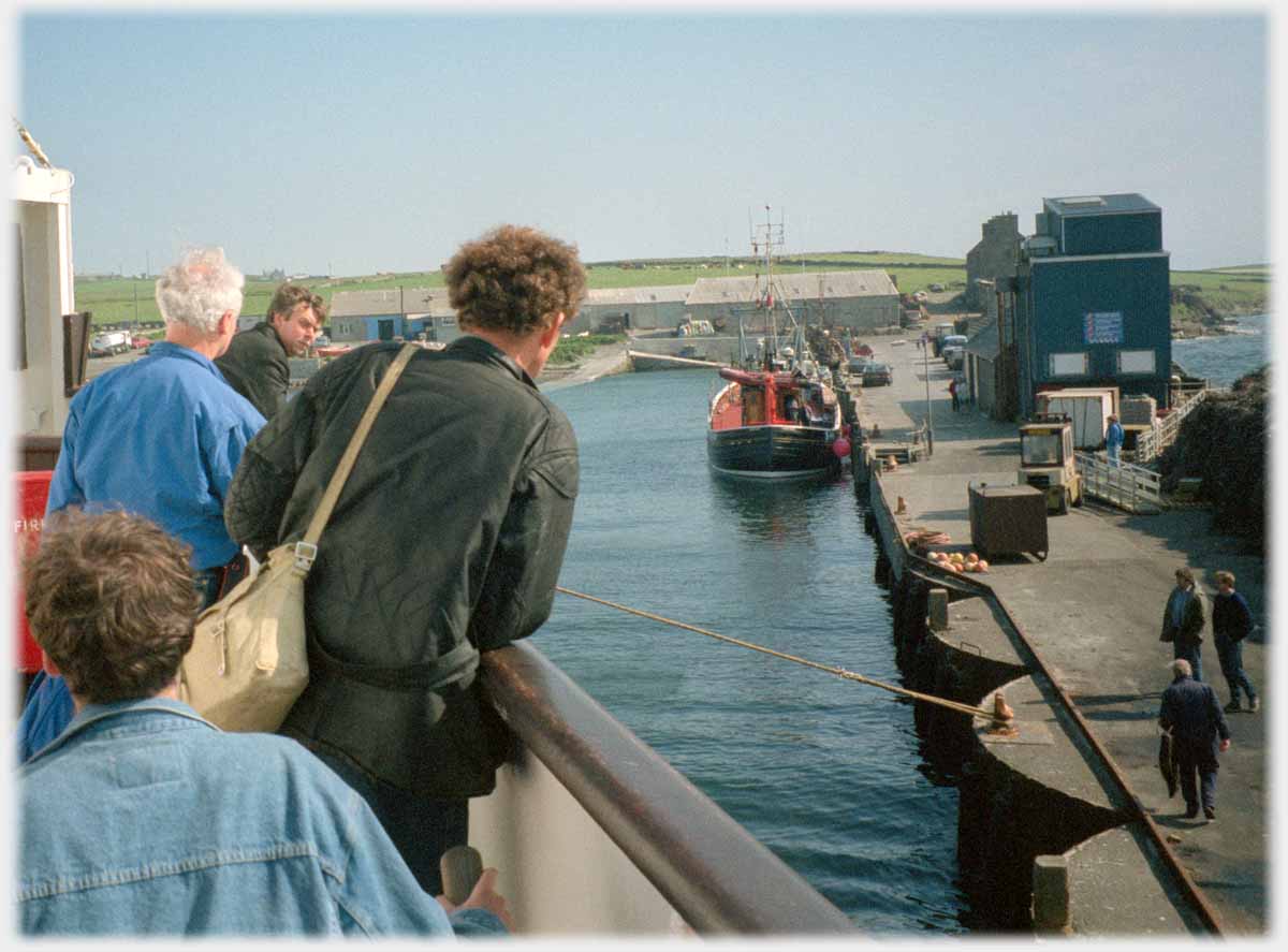 Passengers leaning on rail of ship looking down at quay.