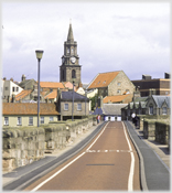 View along narrow bridge with houses and church at far end.