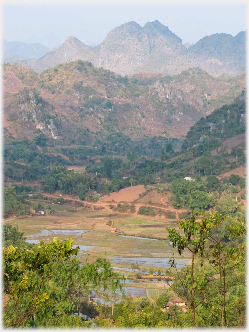 Mountains and hills rising above small wooded valley with houses leading down to river side fields.