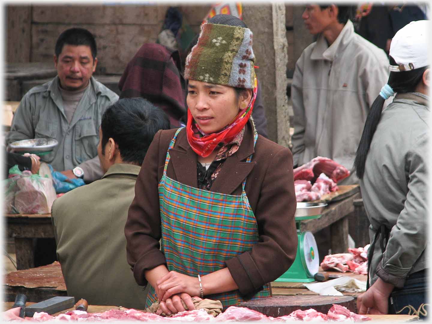 Younger woman standing at meat stall in apron and open topped hat.