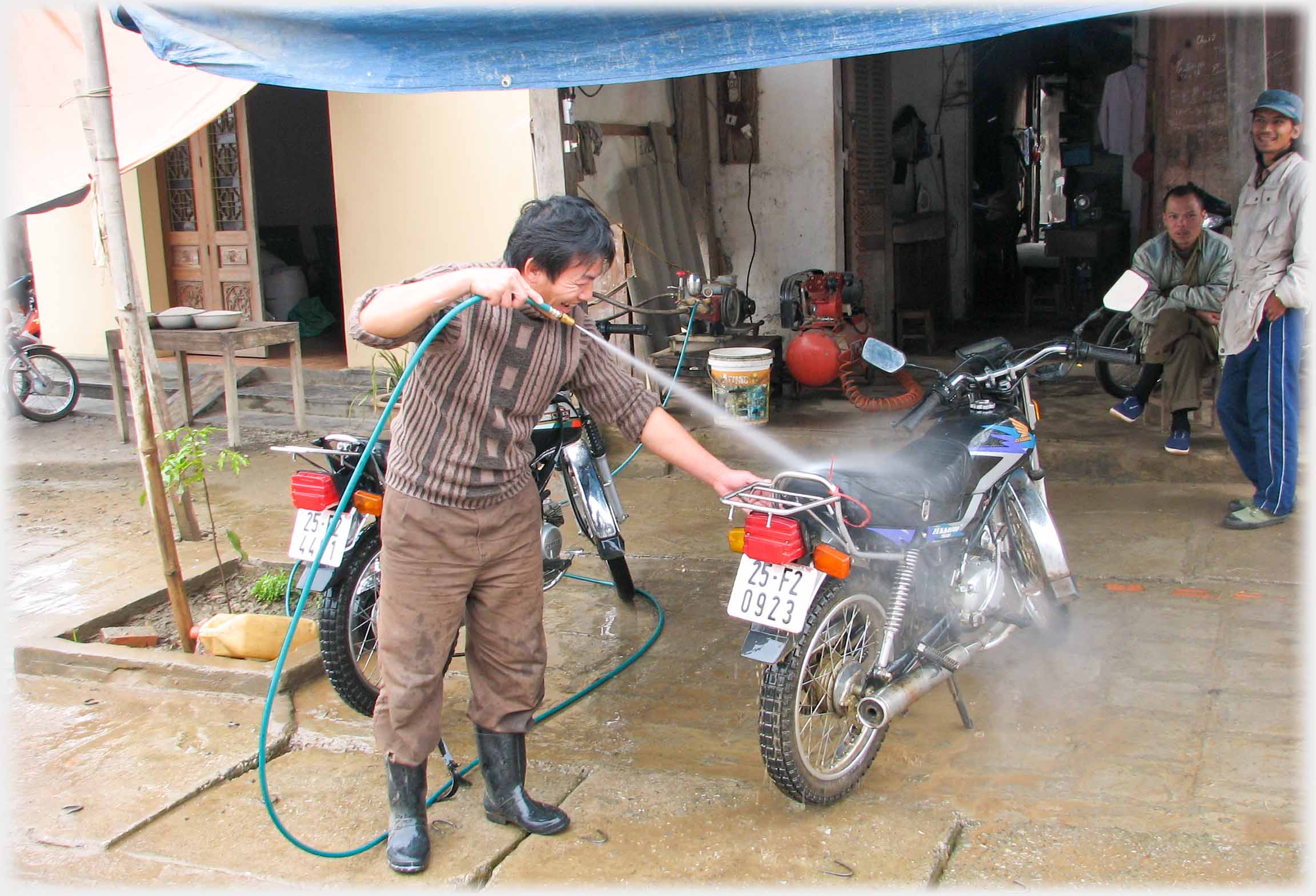 Man with power hose in gum-boots washing motorbike, two men watching.