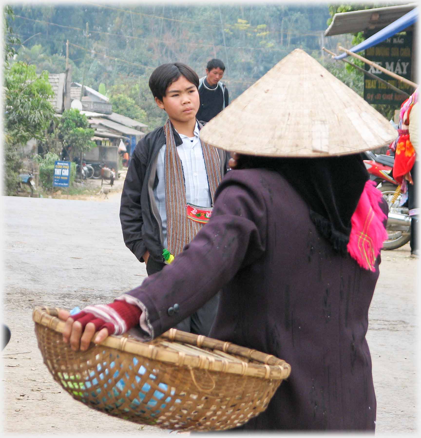 Woman in conical hat holding large flat basket, boy beyond with hair neatly dressed.