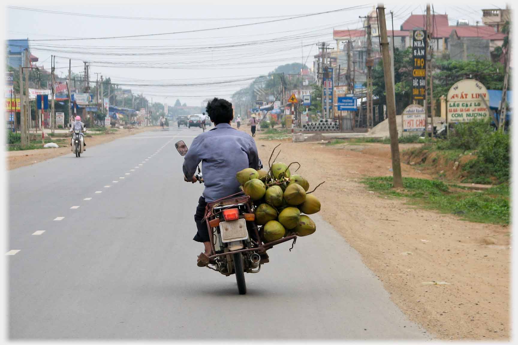 Motorbike being ridden leaning to one side to balance large load of coconuts on other side.