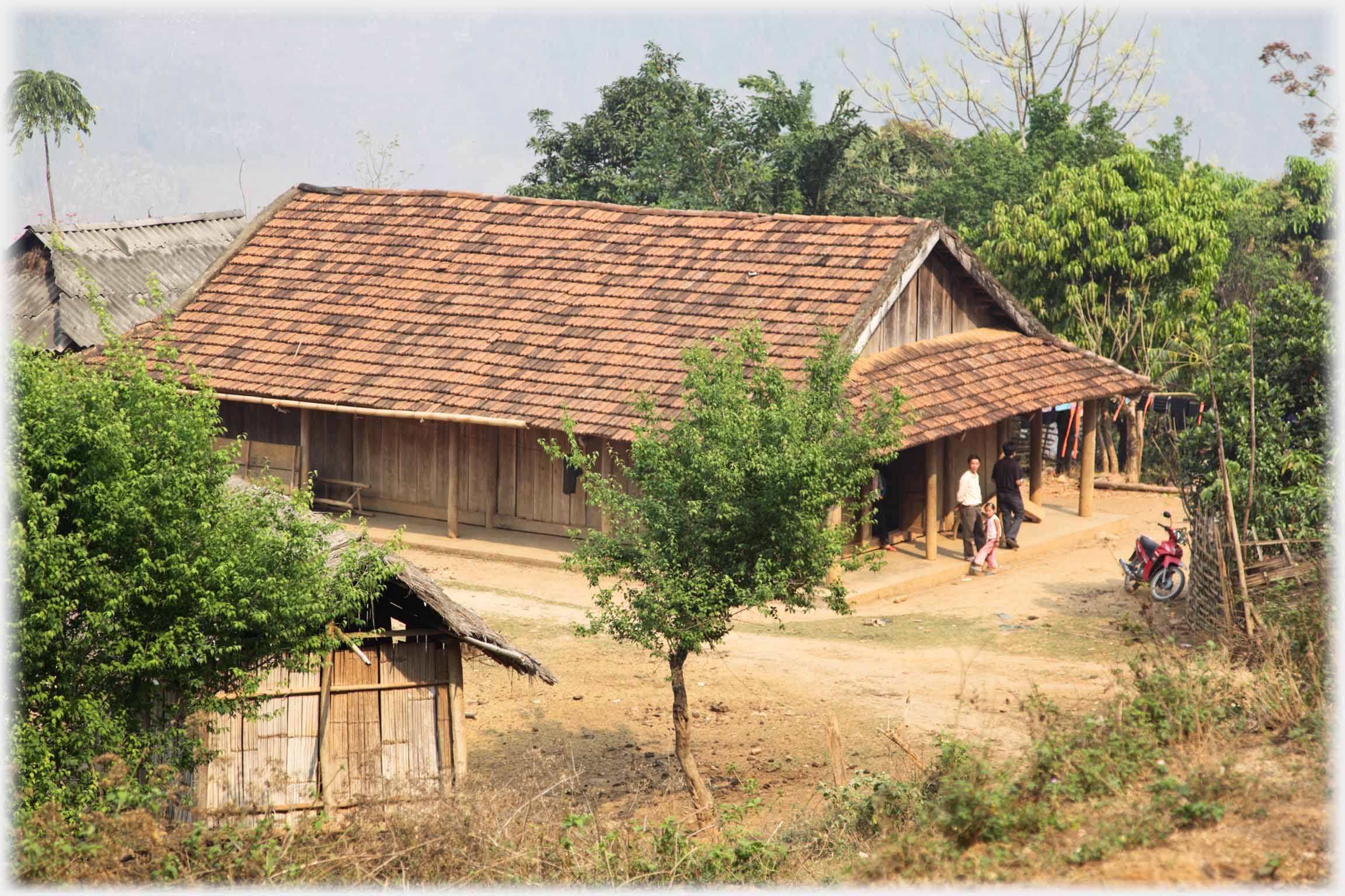 Large pan-tiled house, with roof extending our to cover veranda and there supported on posts. At its rear part of asbestos roof can be seen.