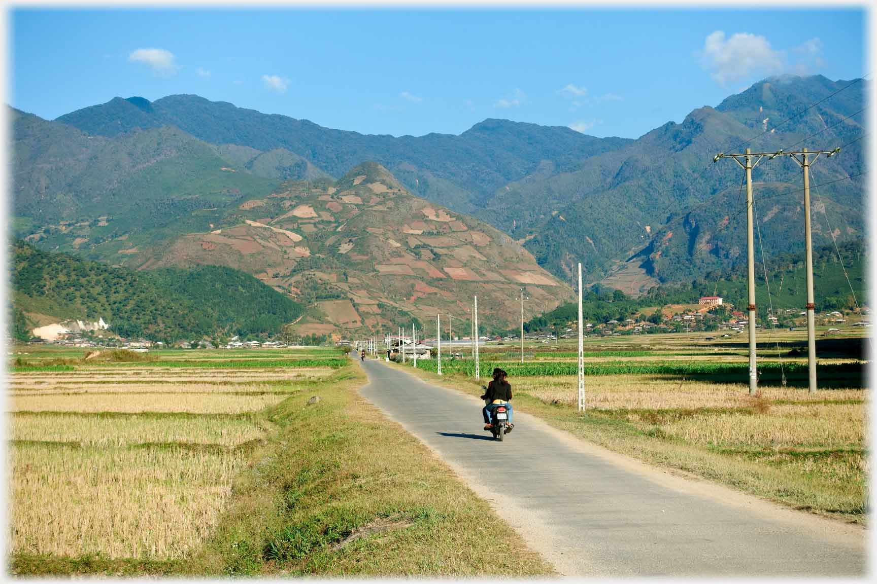 Small straight road leading towards hills, motorcycle on it.