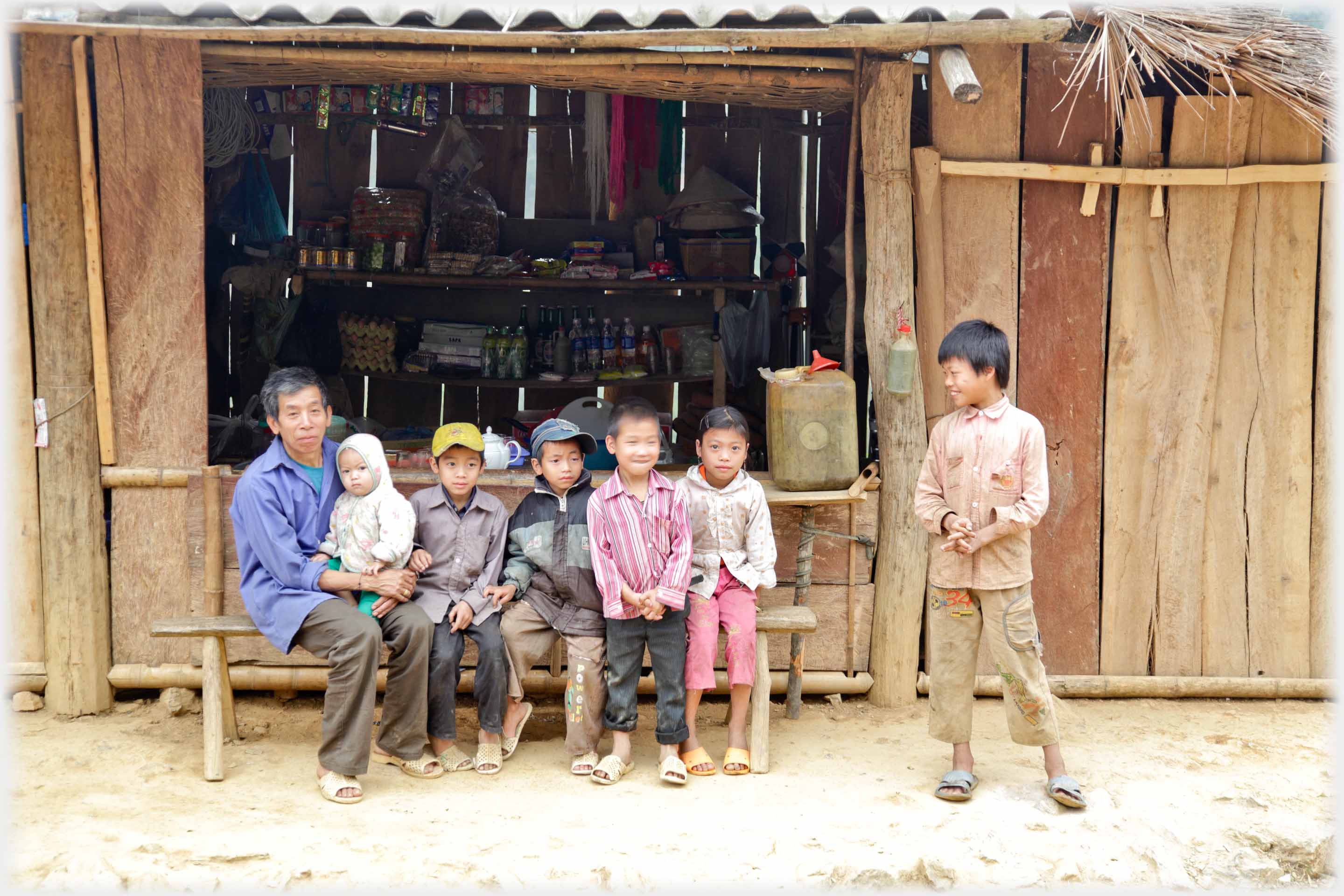 Bench under window of small shop on which a man and five children are sitting, and one older boy standing.