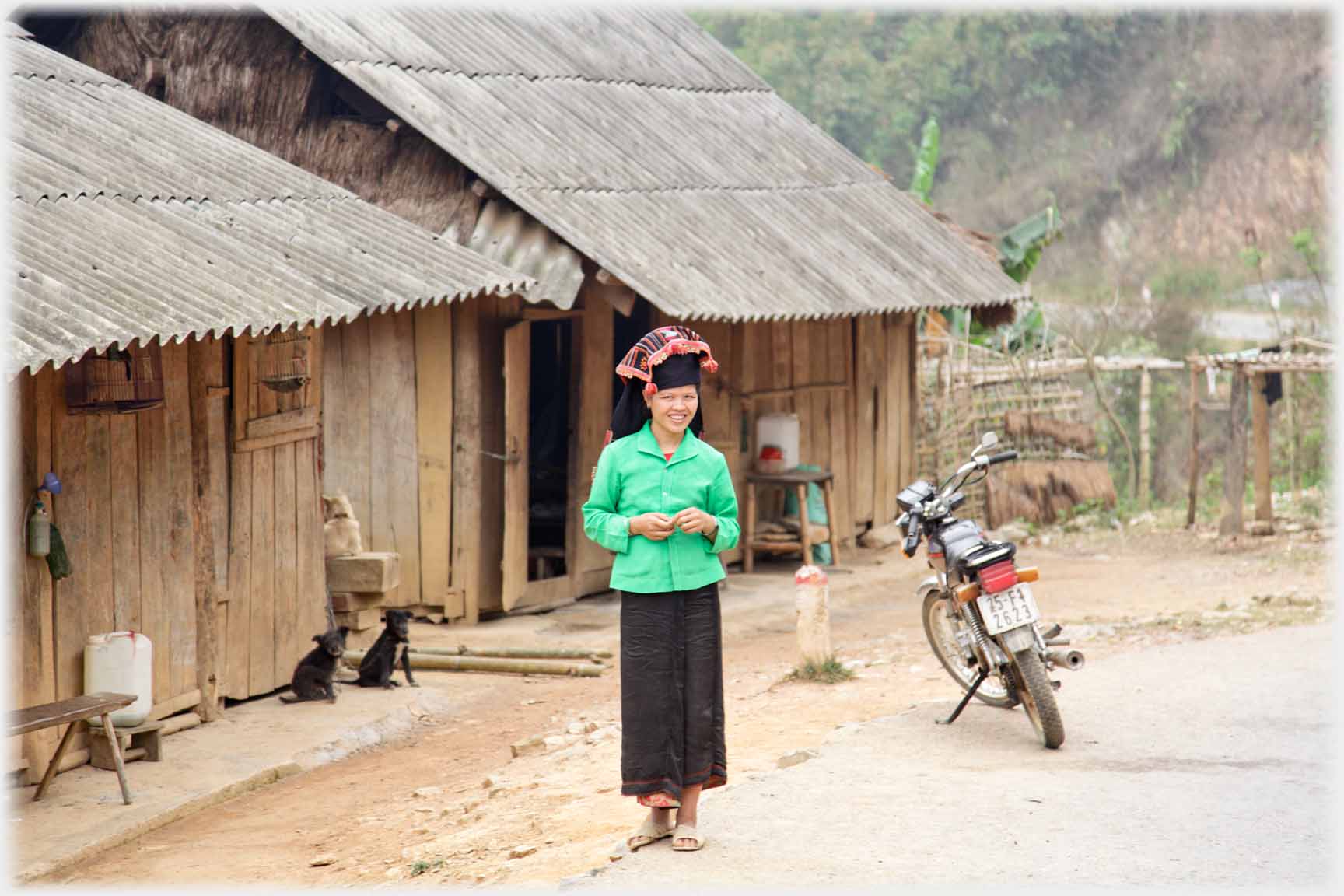Young woman in bright green jacket and Hmung headdress staning in front of houses beside motorbike and two dogs.