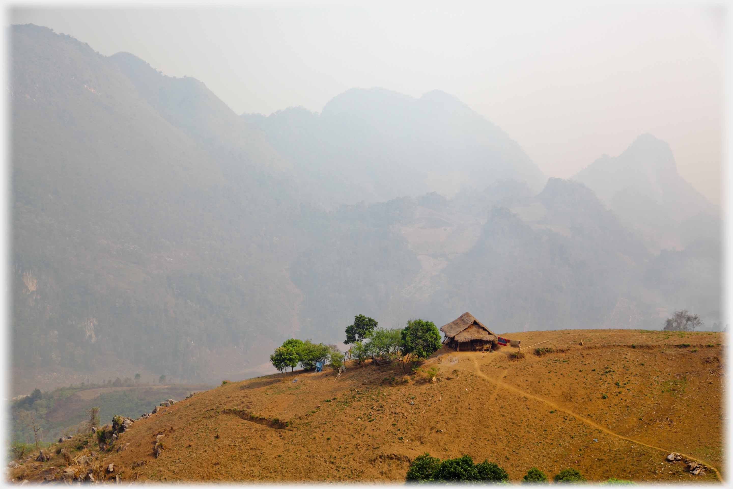 Silhouetted house on hill crest with large mountains looming up through mist beyond.