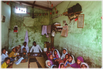 Old school room with two teachers and children sitting on ground.