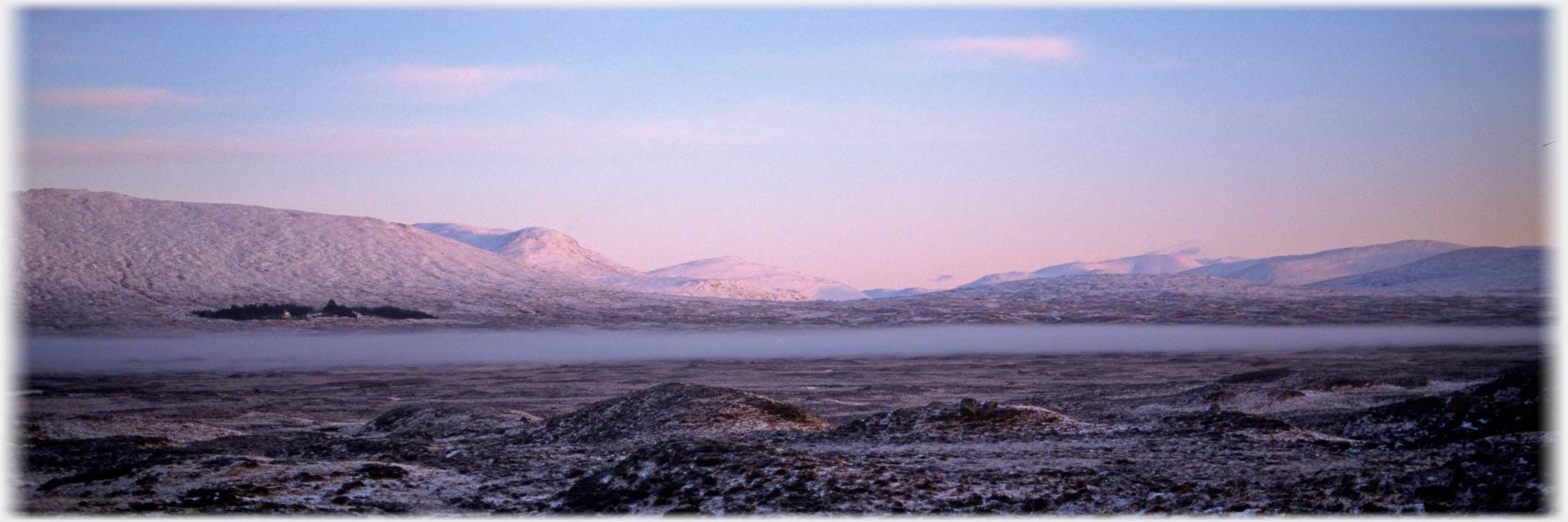 Pink lit snow covered hills across large flat area with mist hanging.