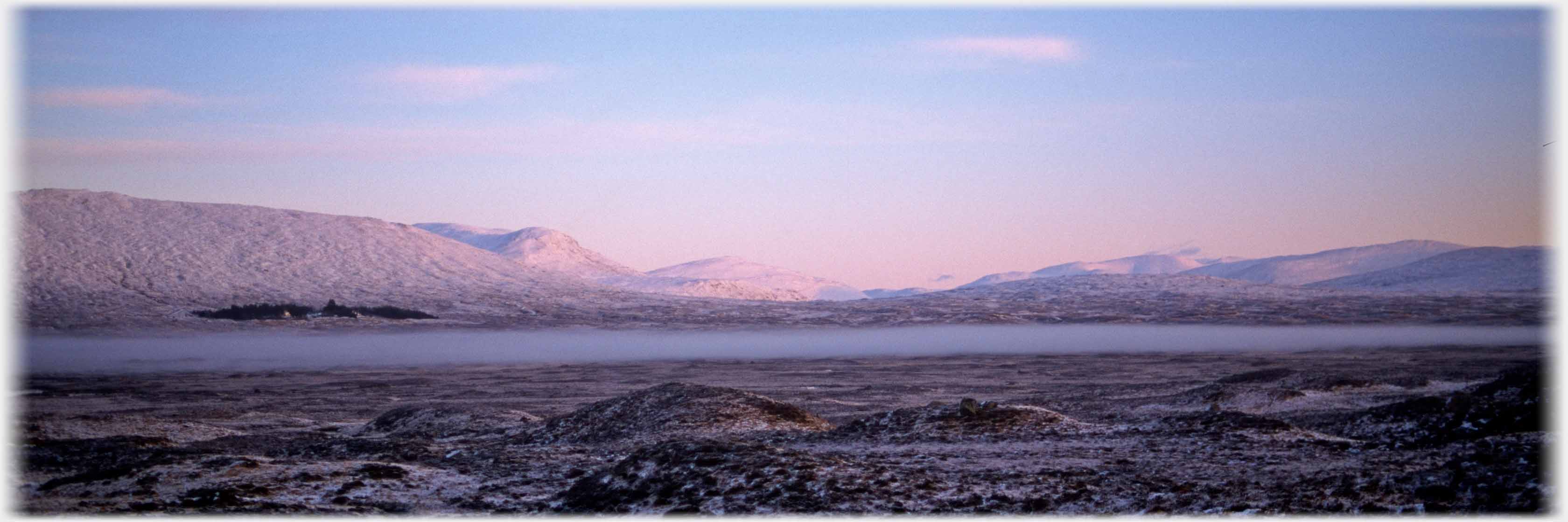 Pink lit snow covered hills across large flat area with mist hanging.