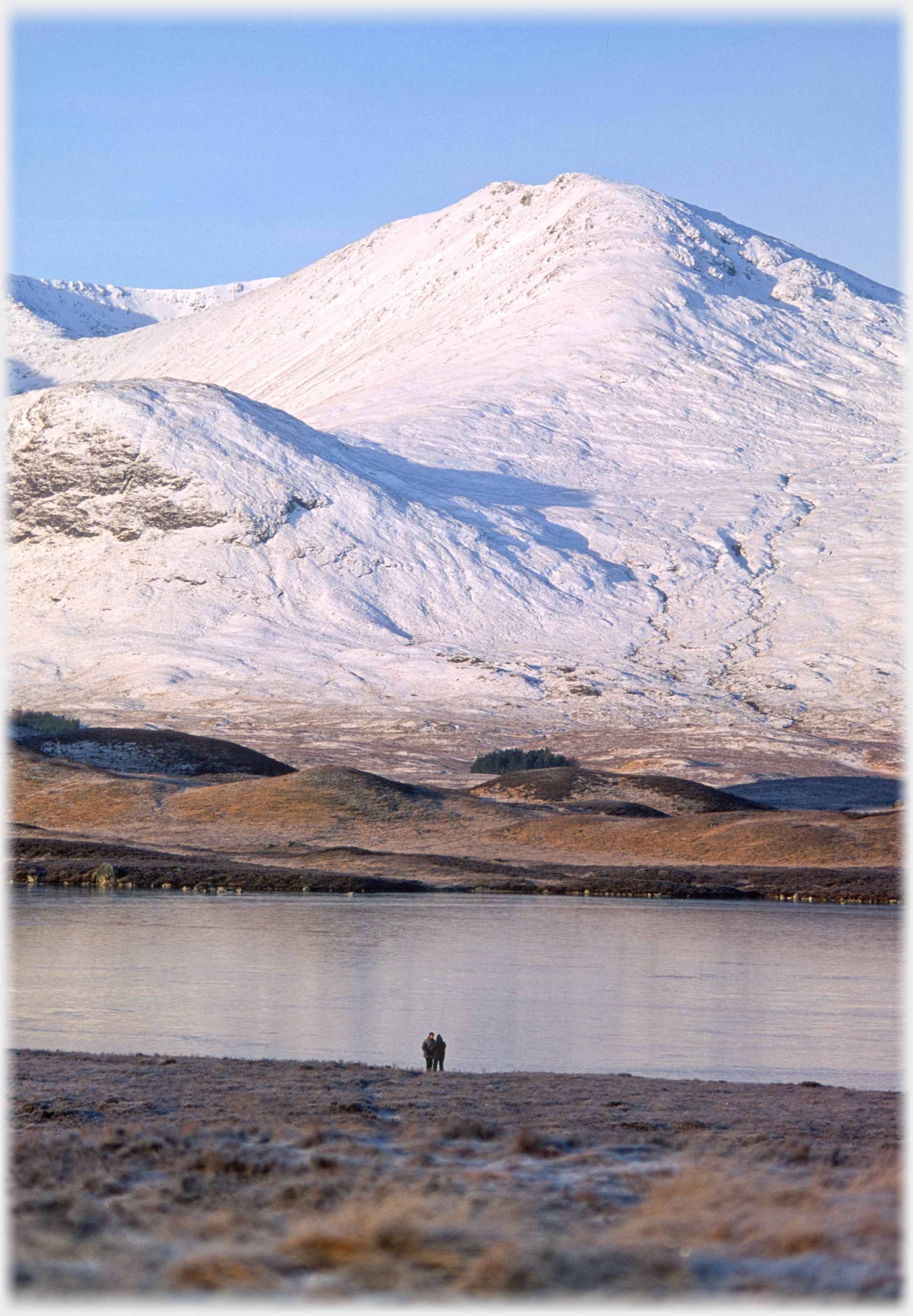 Some distance away two people stand beside a loch with snow covered hill on far side.