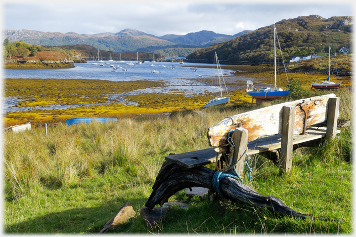 Bench comprised partly of drifwood looking out at low water across boats in bay.