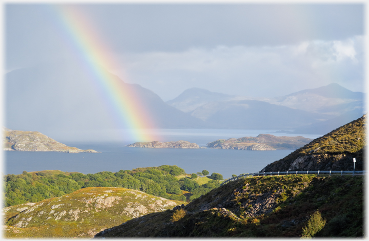 Rainbow touching small island with dark clouds and hills beyond.