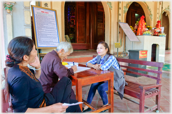 Man writing in donations ledger at table with doner and attendent sitting.