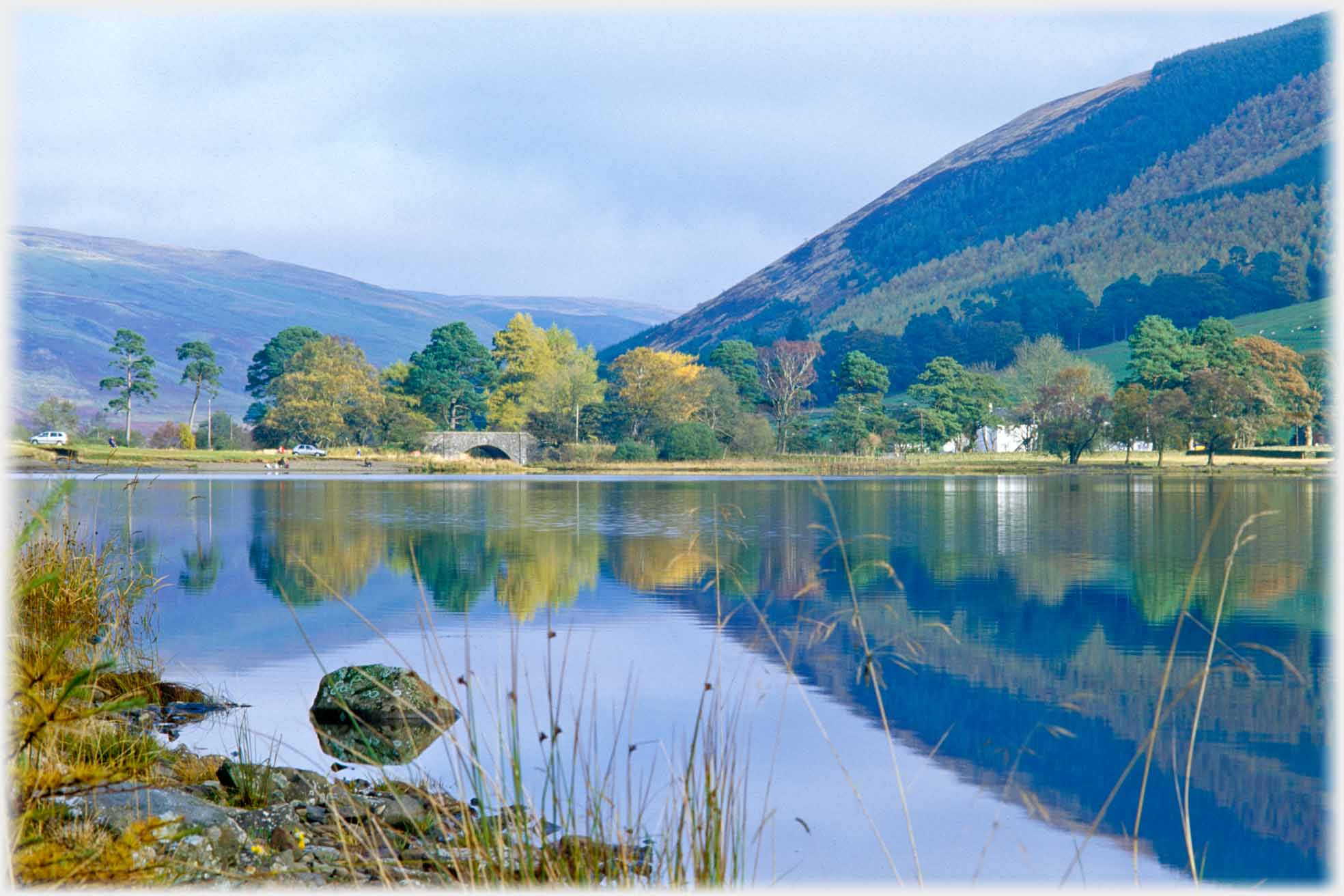 Loch with trees, bridge and hills reflected in it.