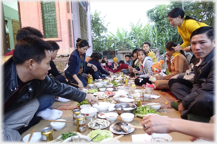 Large group of people sitting on mats on ground with meal.