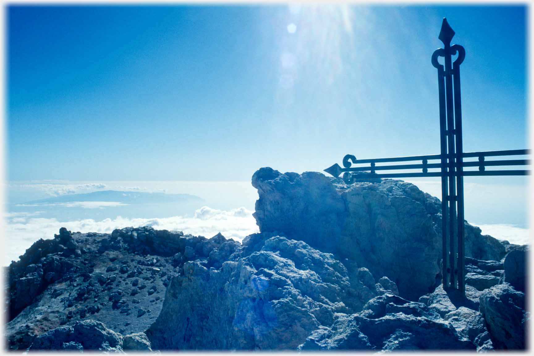 Metal cross, rocks and clouds below.