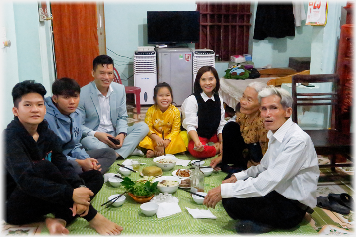 Three generation family group sitting around mat with light meal waiting.