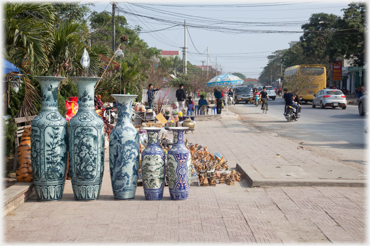Line of five tall vases on pavement by busy road.