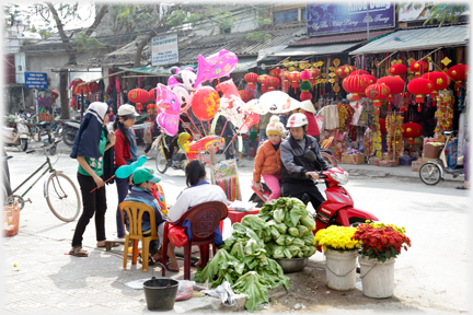 Motorcyclist and passenger inspecting vegetables and flowers at a roadside stall.