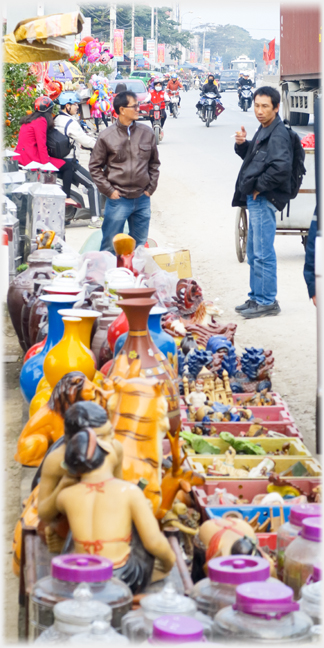 Range of ornimental items with customer and seller standing and traffic just behind them.