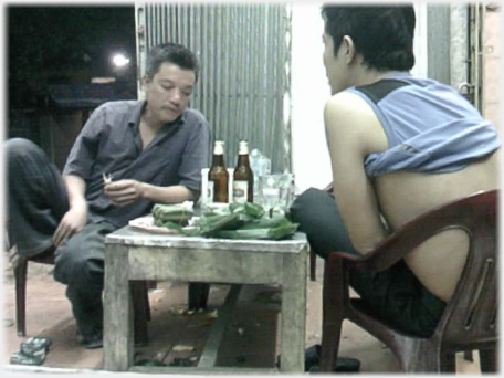 Man feet up on chair and shirt rolled up to oxters, other man leaning on table.