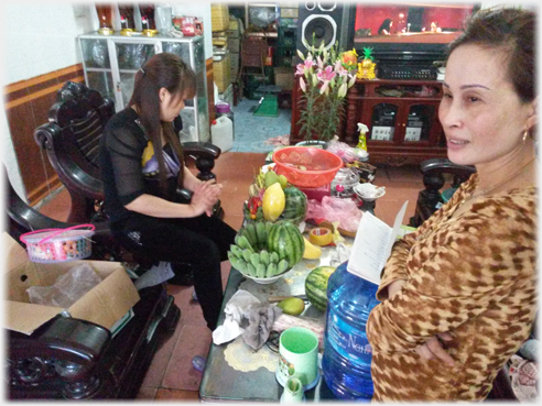 Table laden with fruits, formal chairs, woman stranding looking, daughter sitting.