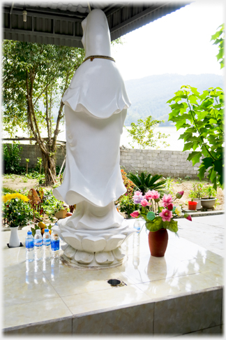 Rear of Standing white Buddha with lake just visible over wall beyond.