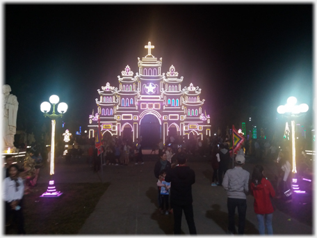 Entrance lamps, statue and crowd in front of church facade.