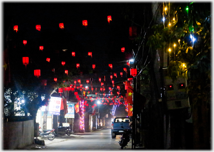 Street with small red lanterns hanging over it off into the distance, parked car, lit shop.