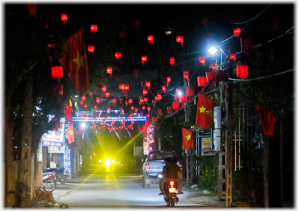 Street with small red lanterns hanging over it off into the distance, motorbikes passing.