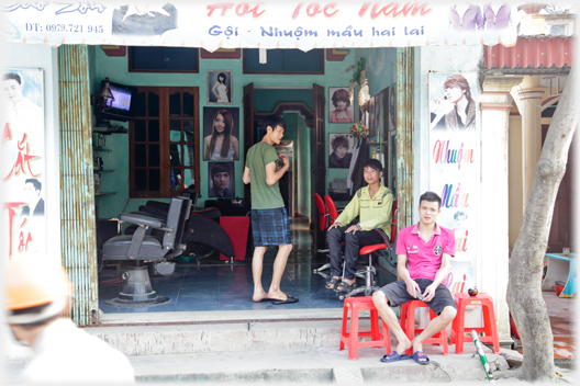 Three young men sit at the entrance to a barbers shop.