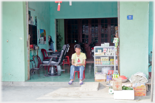 A man sits at the entrance to a hairdresser beside a cabinet of soft drinks.