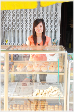 A girl standing behind a cabinet of cakes.