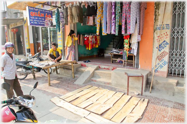 Clothes shop with man standing ourside and another squatting on a table laying out shirts.