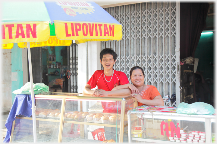 A mother links arms with her  much taller son behind a cabinet of cakes.