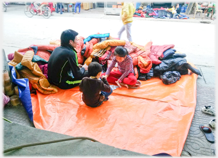 A man sits at the roadside on a tarpaulin beside two children who are playing cards behind a pile of padded winter jackets.