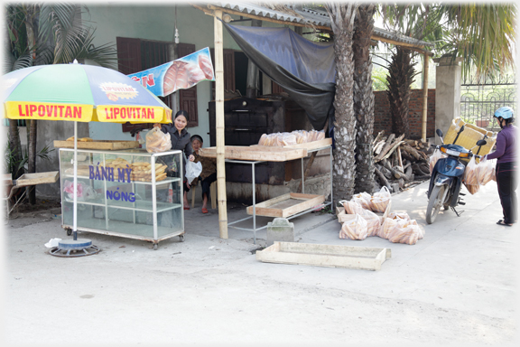 Woman behind cabinet with breads, piles of bread to one side, oven behind and delivery motorcyclist loading up.