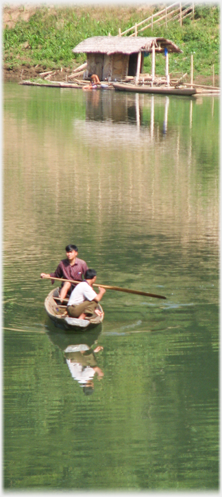 Two men in small open boat on river with covered boat beyond by the shore.