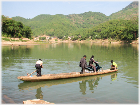 Narrow open boat being paddled with three passengers, one sitting on a motorbike.